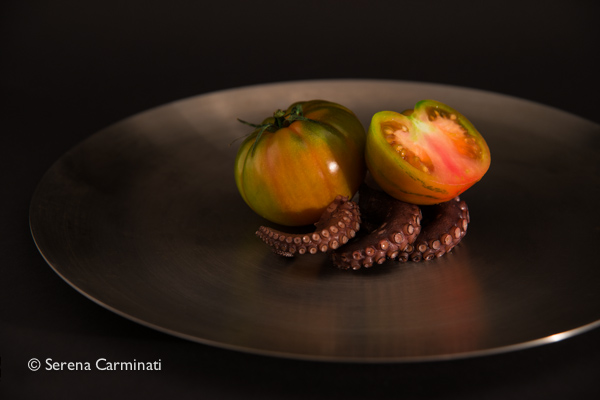 Green tomatoes on a few octopus tentacles on a bronze plate with a black paper background. The plate is cut out of the frame on  the left hand side.